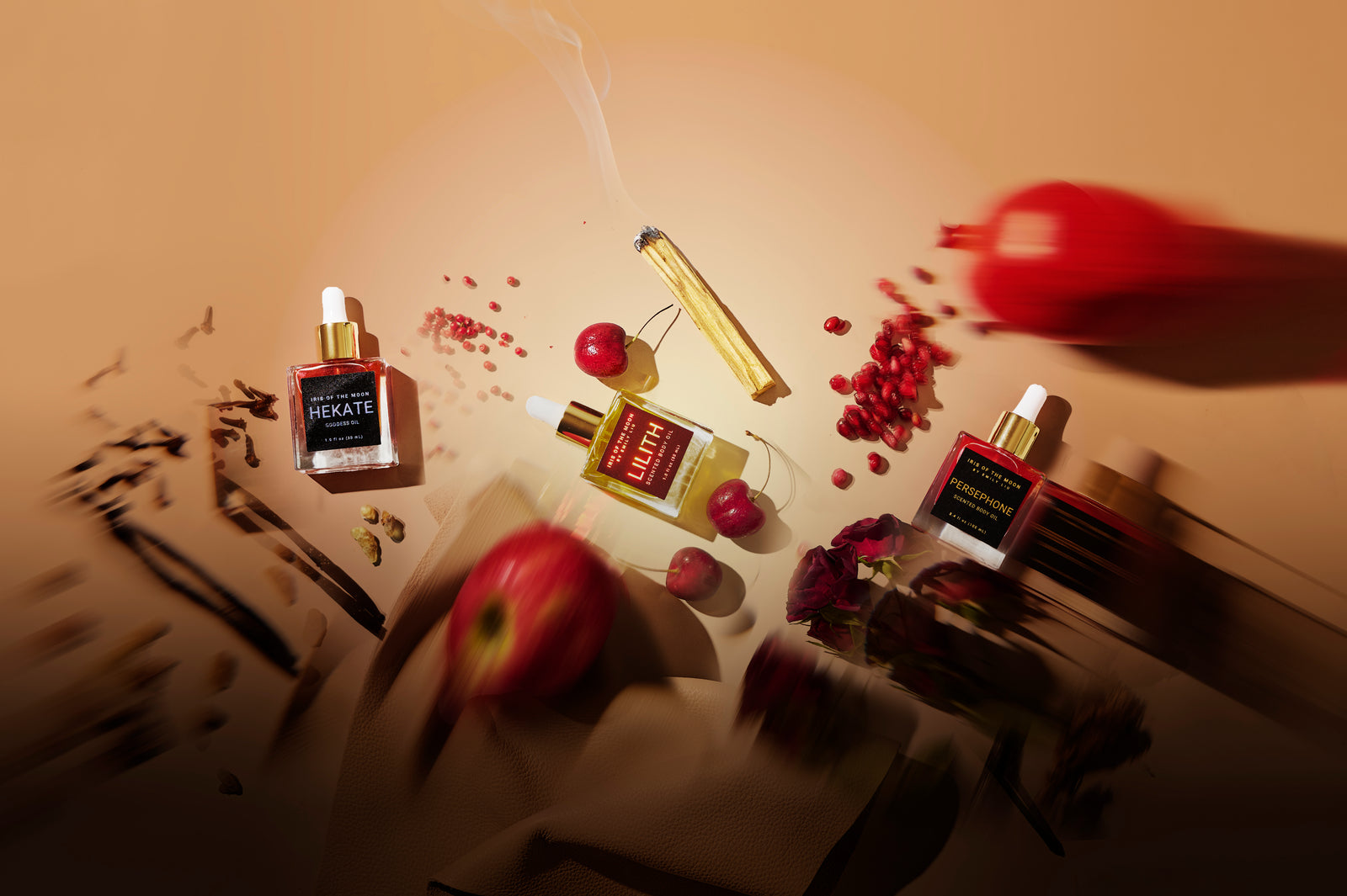 An artistic top-down shot features three "Iris of the Moon" oil bottles surrounded by cherries, pomegranate seeds, a smoking palo santo stick, and blurred botanical elements on a warm tan surface.