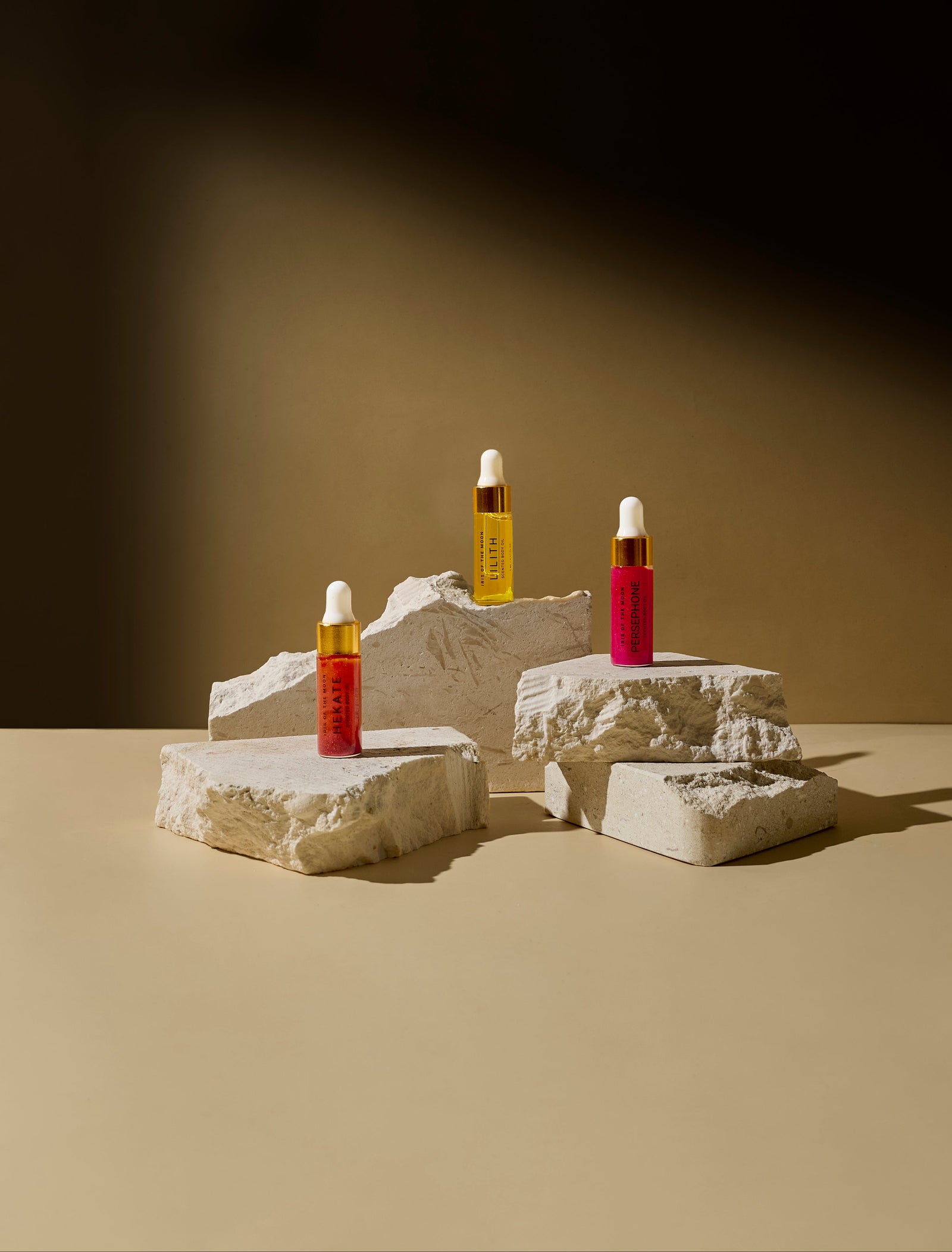 Three small, labeled elixir dropper bottles in red, amber, and pink stand on stacked, rough-hewn stone pedestals against a warm, minimalist backdrop.