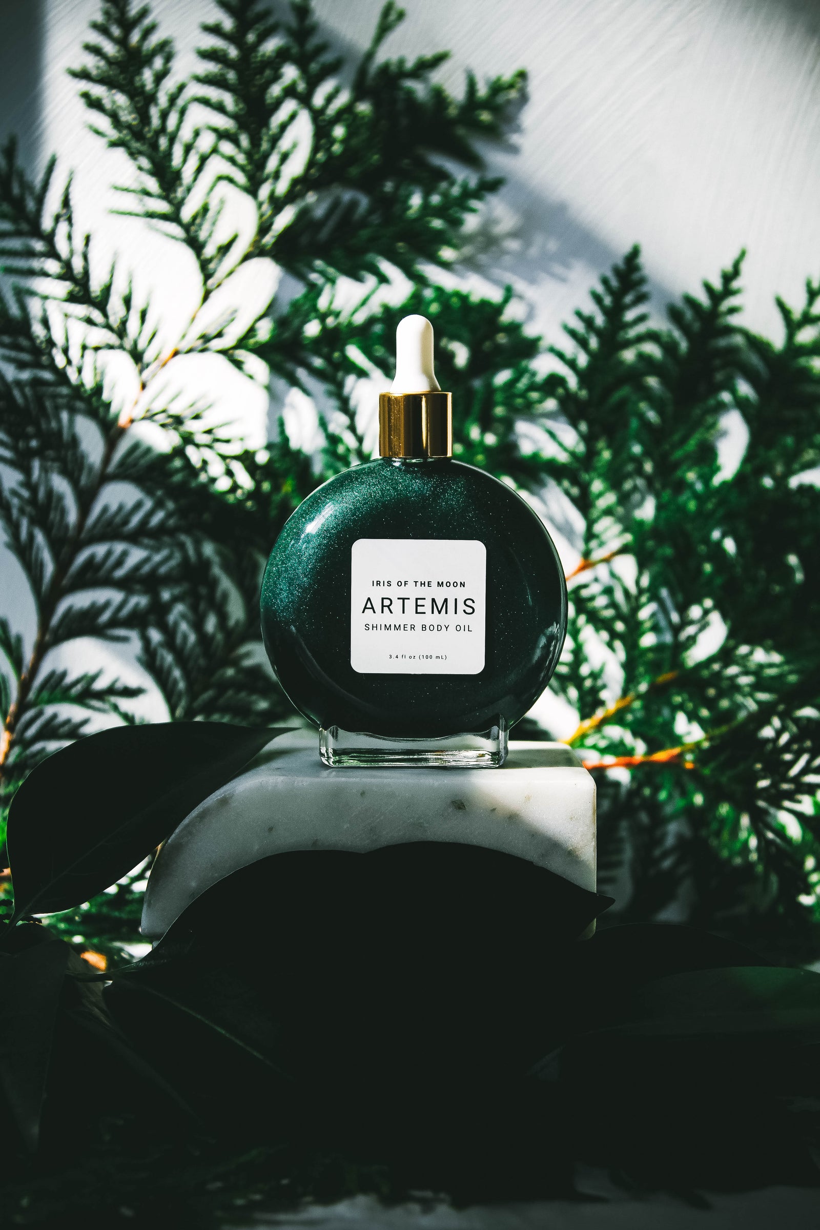 Bottle of Artemis on a marble stand against a backdrop filled with dark green leaves and ferns.