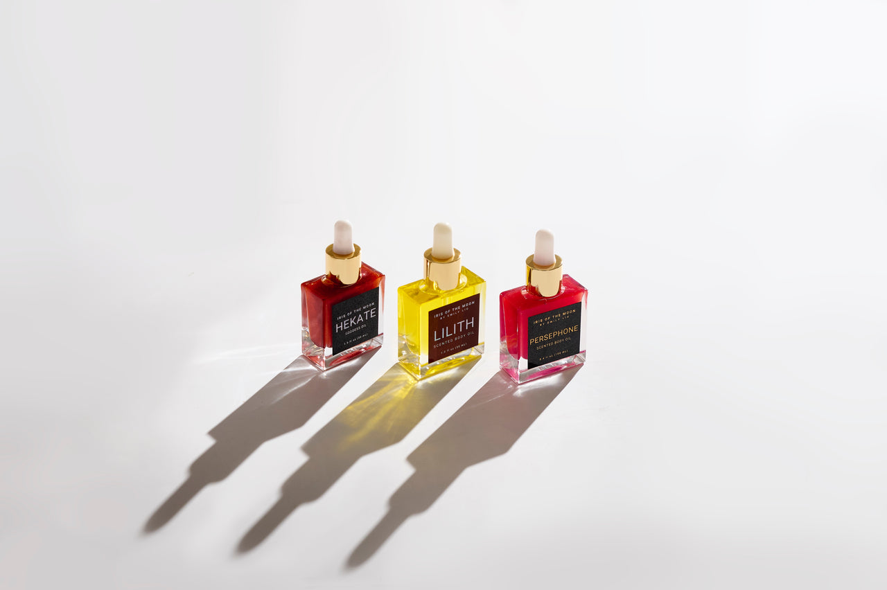 three square bottles of Iris of the Moon body oils—Hekate, Lilith, and Persephone—on a white background with dark shadows at a slant.