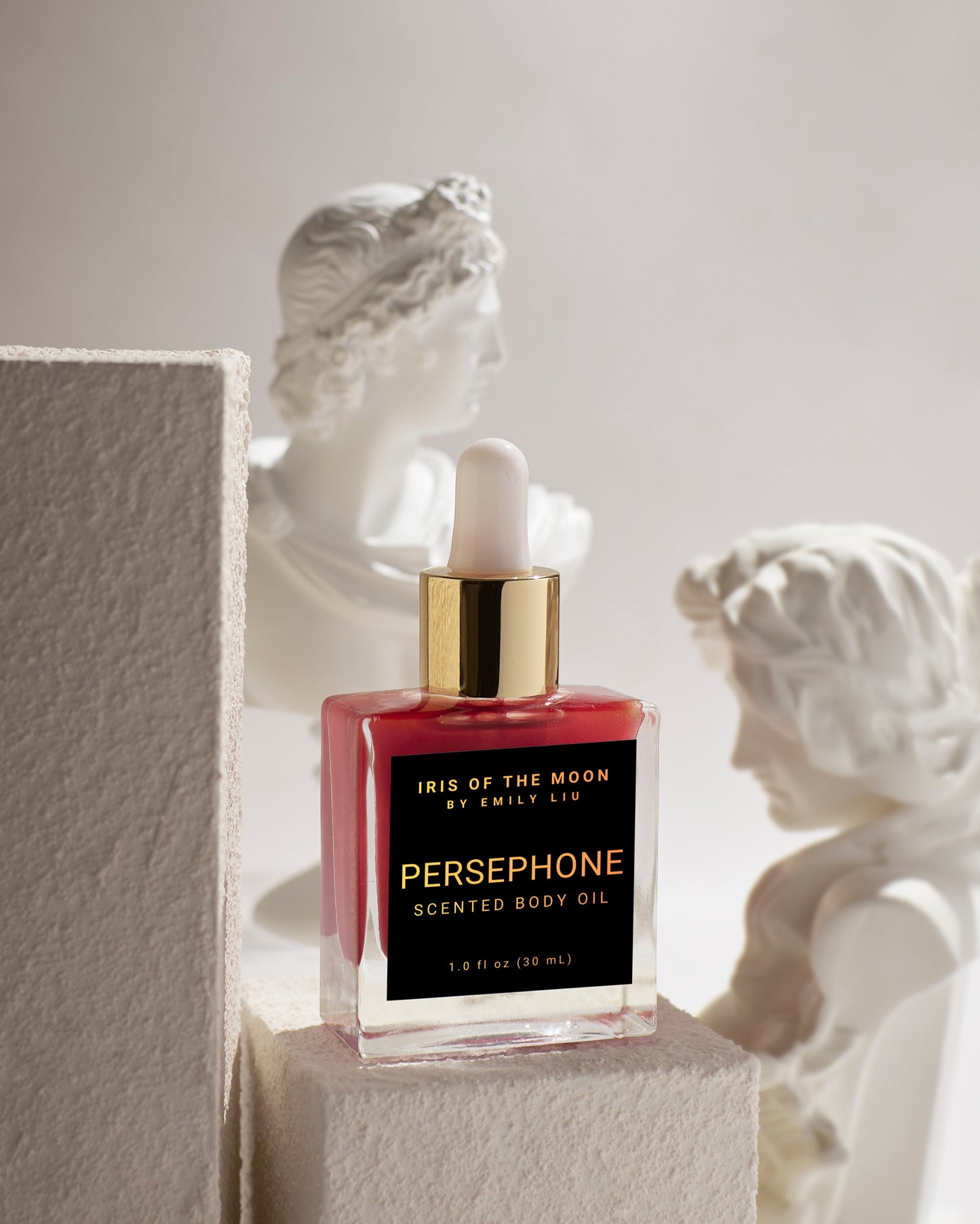 Persephone scented body oil bottle with gold cap on a pedestal in front of classical sculptures.