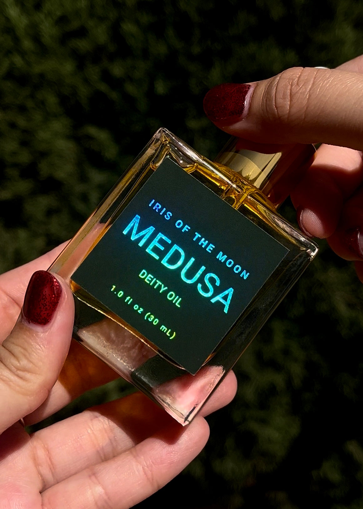 Bottle of 'Iris of the Moon Medusa Deity Oil' held by a person with red nail polish against a dark background.